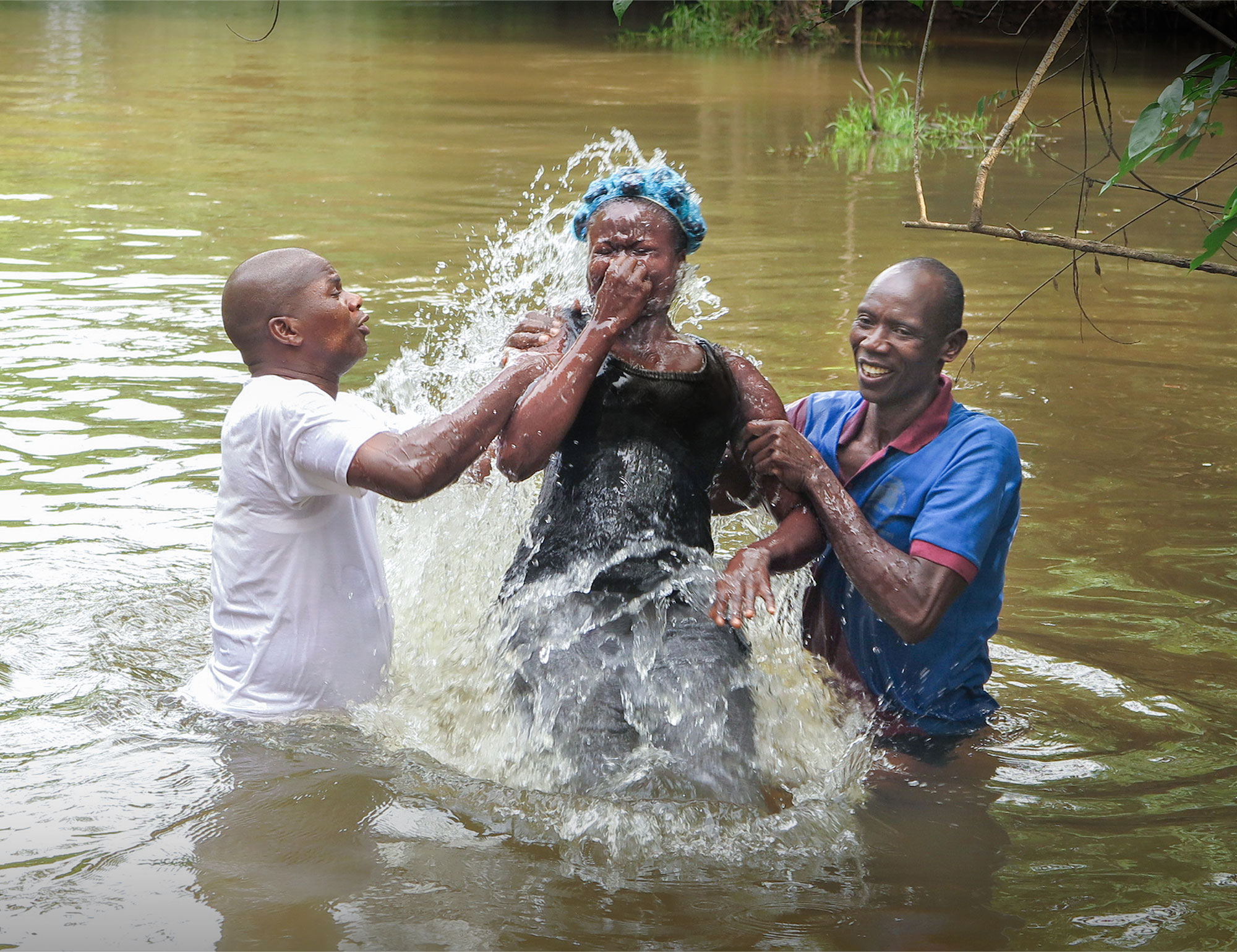 Dos hombres ayudan a una mujer a ser bautizada en un cuerpo de agua.