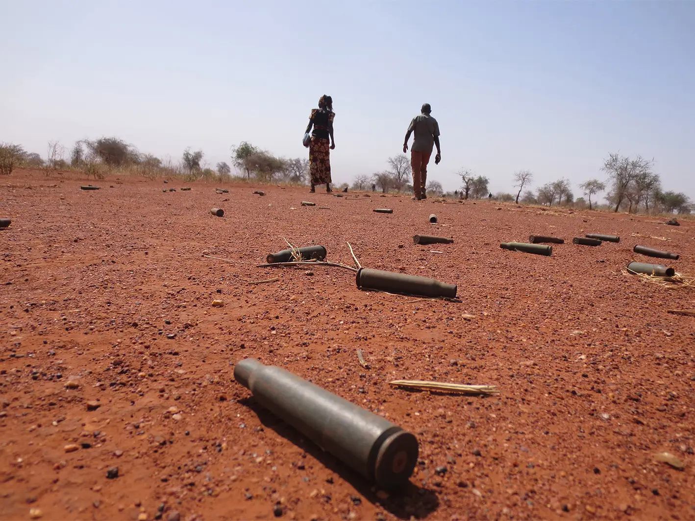 Two people walking through red dirt field that is scattered with bullet casings