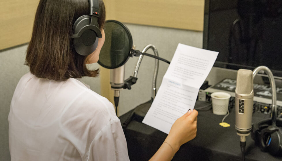 Woman with headphones on reading a piece of paper into a microphone