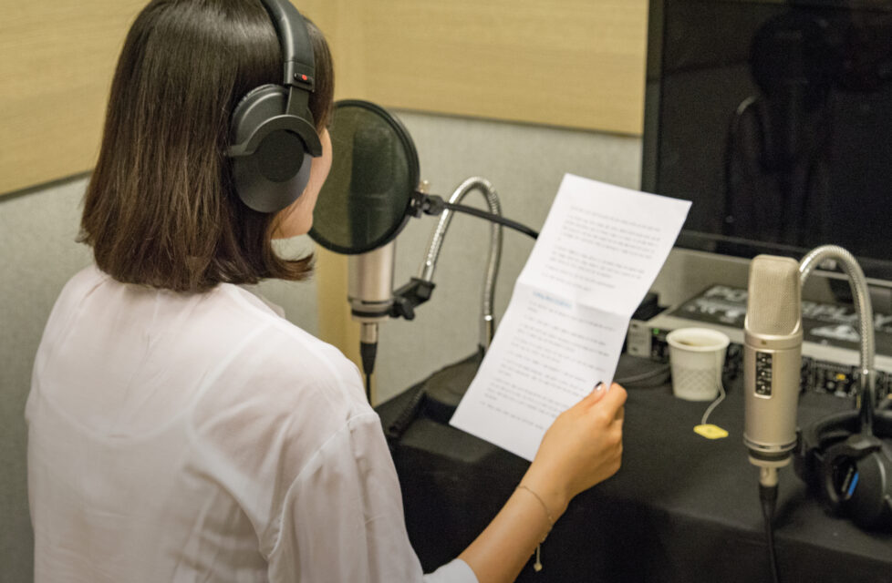 Woman with headphones on reading a piece of paper into a microphone