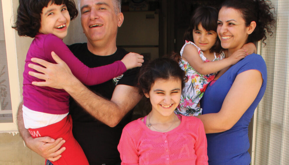 a husband and wife stand with their three girls outside their house smiling