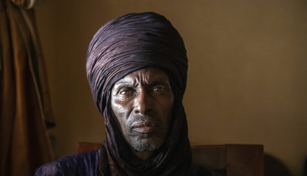 A Fulani man wearing a traditional head covering looks into the camera